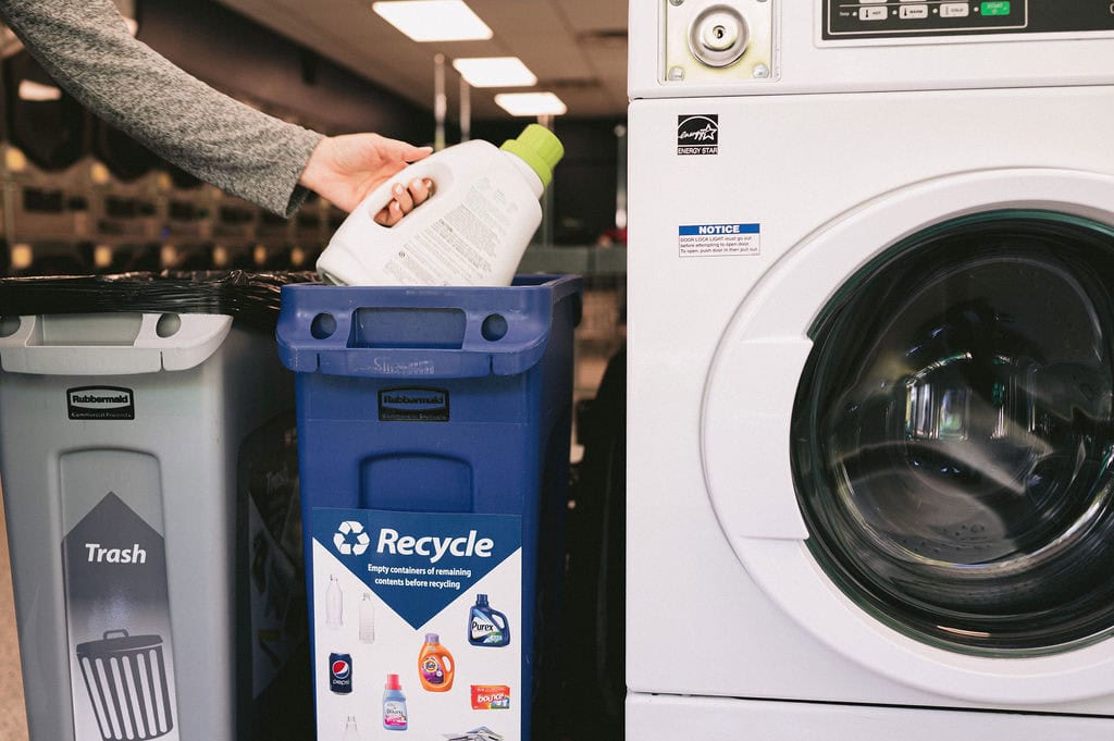 A trash bin, recycling bin, and washing machine sit in a row in a laundromat. An arm reaches to put a detergent bottle in the recycling bin.
